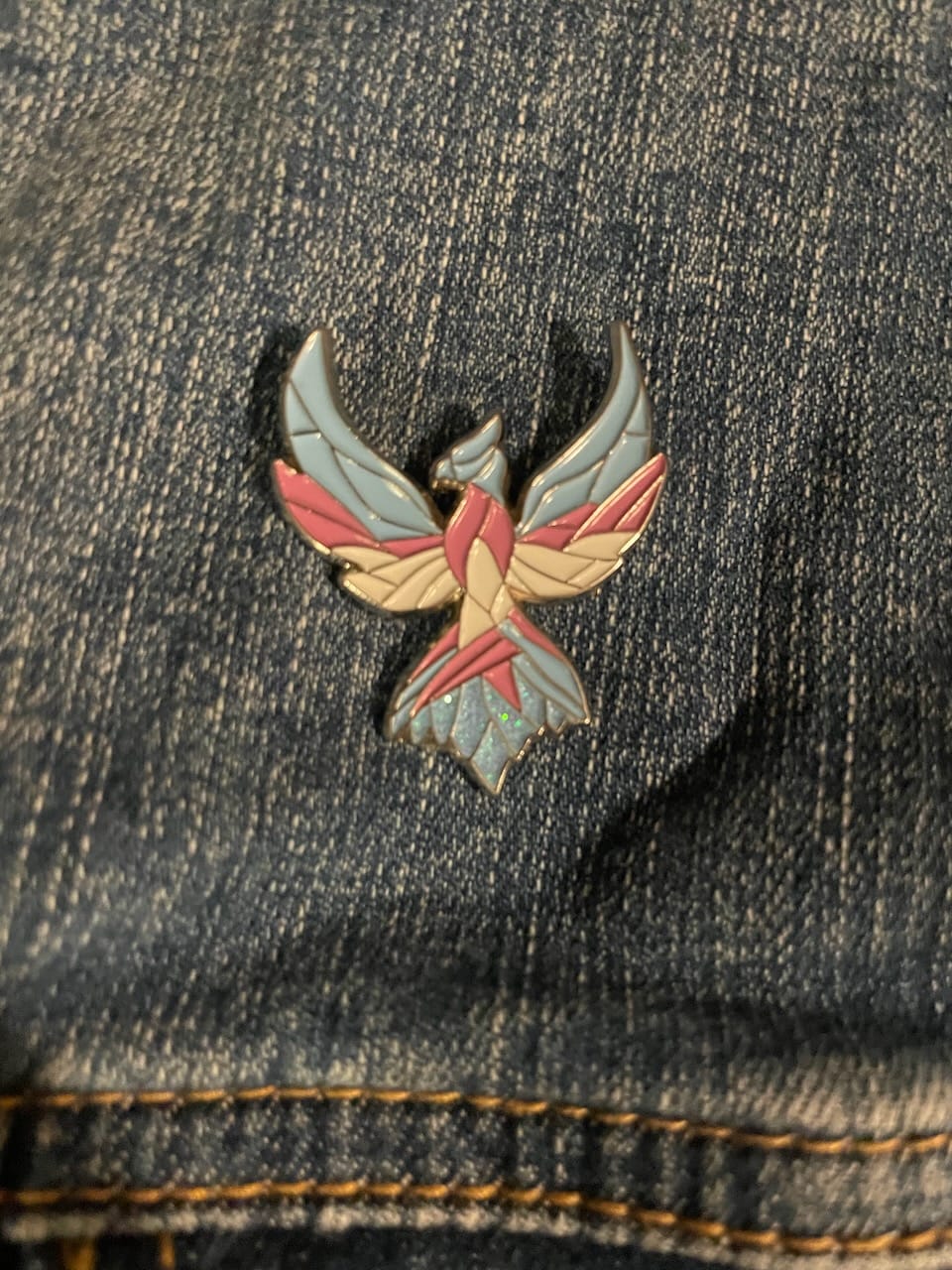 An enamel pin of a phoenix, painted in trans colors, pinned on a denim jacket.