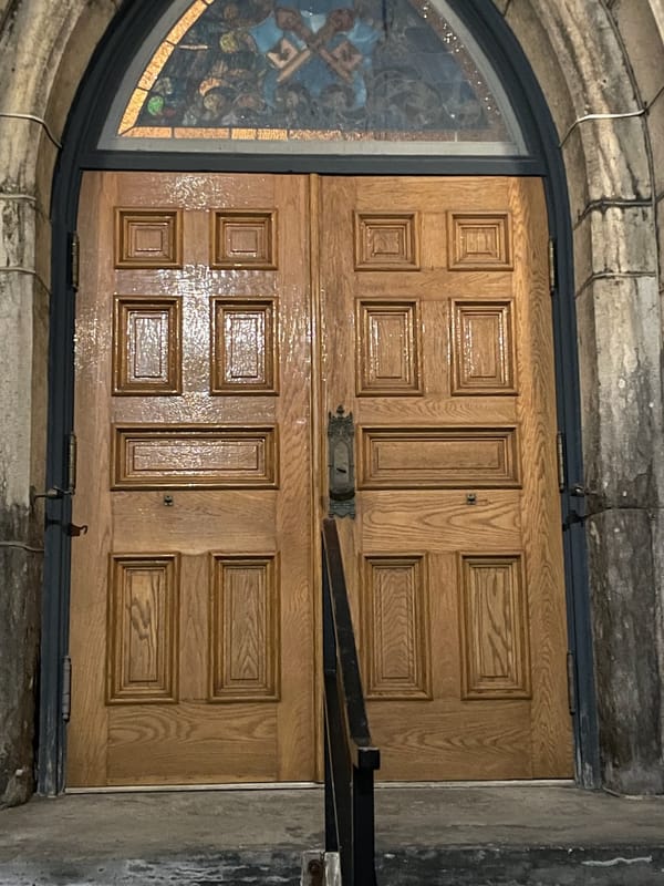 The wooden double-doors of a church.  A stained-glass window is above the doors.